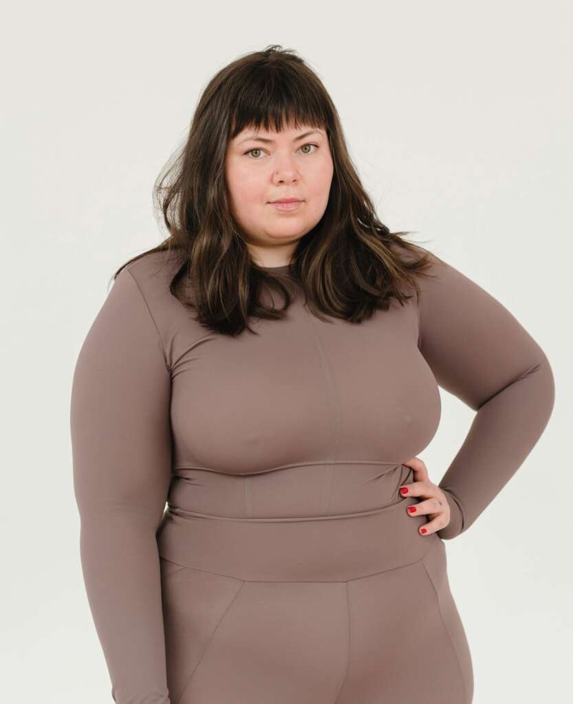 Overweight white, brunette woman standing in a skintight brown outfit with her arm on her hip.